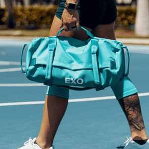 A person carries an aqua-blue EXO sports duffel bag while walking on a basketball court, wearing a smartwatch and shorts, with a tattoo on their calf.