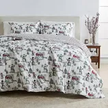 Holiday-themed bedding set featuring a comforter and pillowcases with snowmen, trees, and red barns, complemented by a striped reverse side. A simple nightstand with decorative branches and candles is visible.