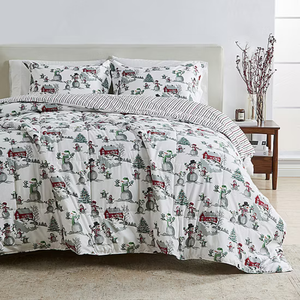 Holiday-themed bedding set featuring a comforter and pillowcases with snowmen, trees, and red barns, complemented by a striped reverse side. A simple nightstand with decorative branches and candles is visible.