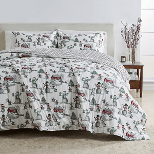 Holiday-themed bedding set featuring a comforter and pillowcases with snowmen, trees, and red barns, complemented by a striped reverse side. A simple nightstand with decorative branches and candles is visible.