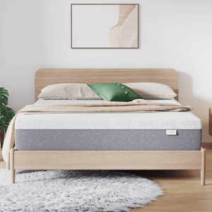 A modern bedroom features a wooden bed frame with a neatly made bed, showcasing a mattress. The setup includes beige and green pillows, a beige blanket, and a fluffy gray rug on a wooden floor, complemented by minimalistic wall art and a plant.