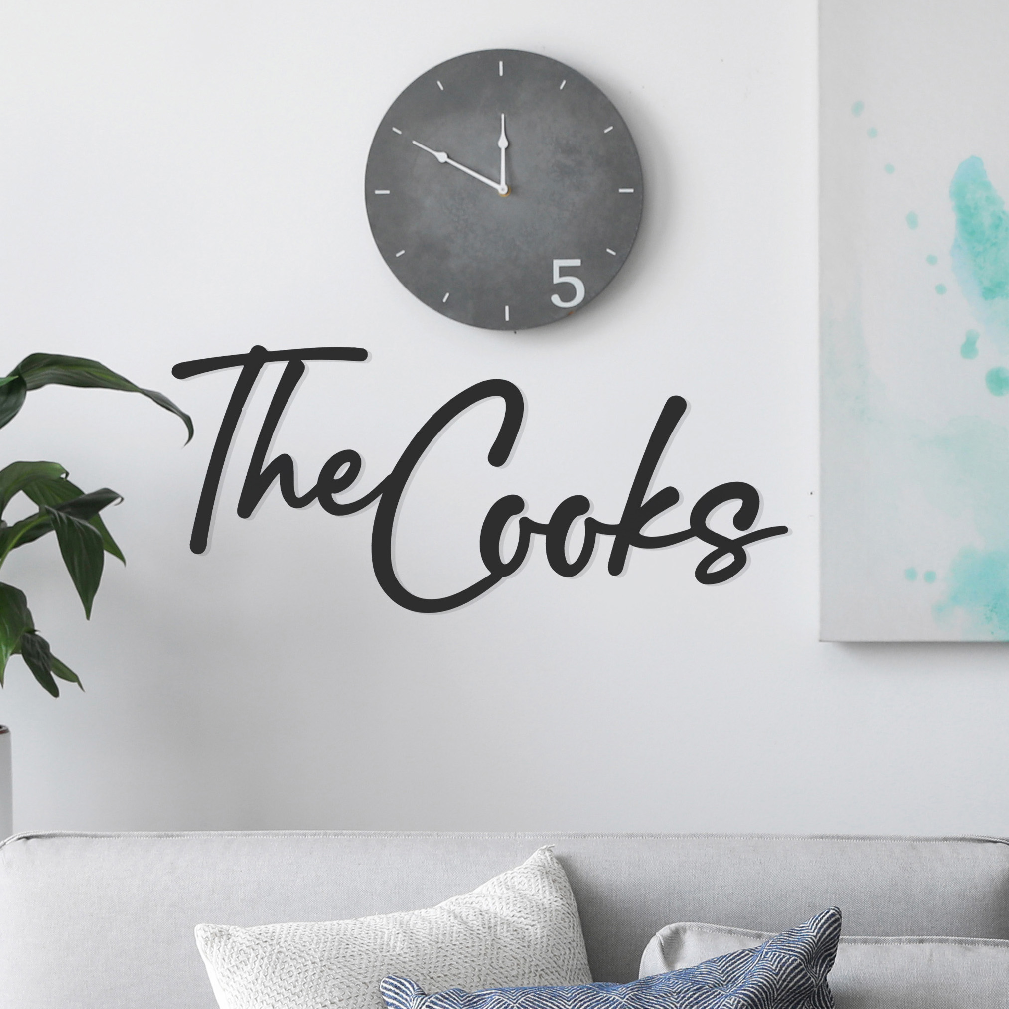 Wall decal reading \“The Cooks\“ in cursive, placed above a sofa with cushions and next to a houseplant, accompanied by a round gray clock displaying 1:50.