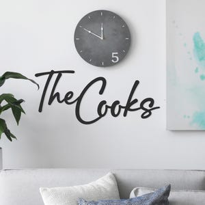 Wall decal reading \“The Cooks\“ in cursive, placed above a sofa with cushions and next to a houseplant, accompanied by a round gray clock displaying 1:50.