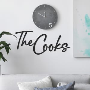 Wall decal reading \“The Cooks\“ in cursive, placed above a sofa with cushions and next to a houseplant, accompanied by a round gray clock displaying 1:50.