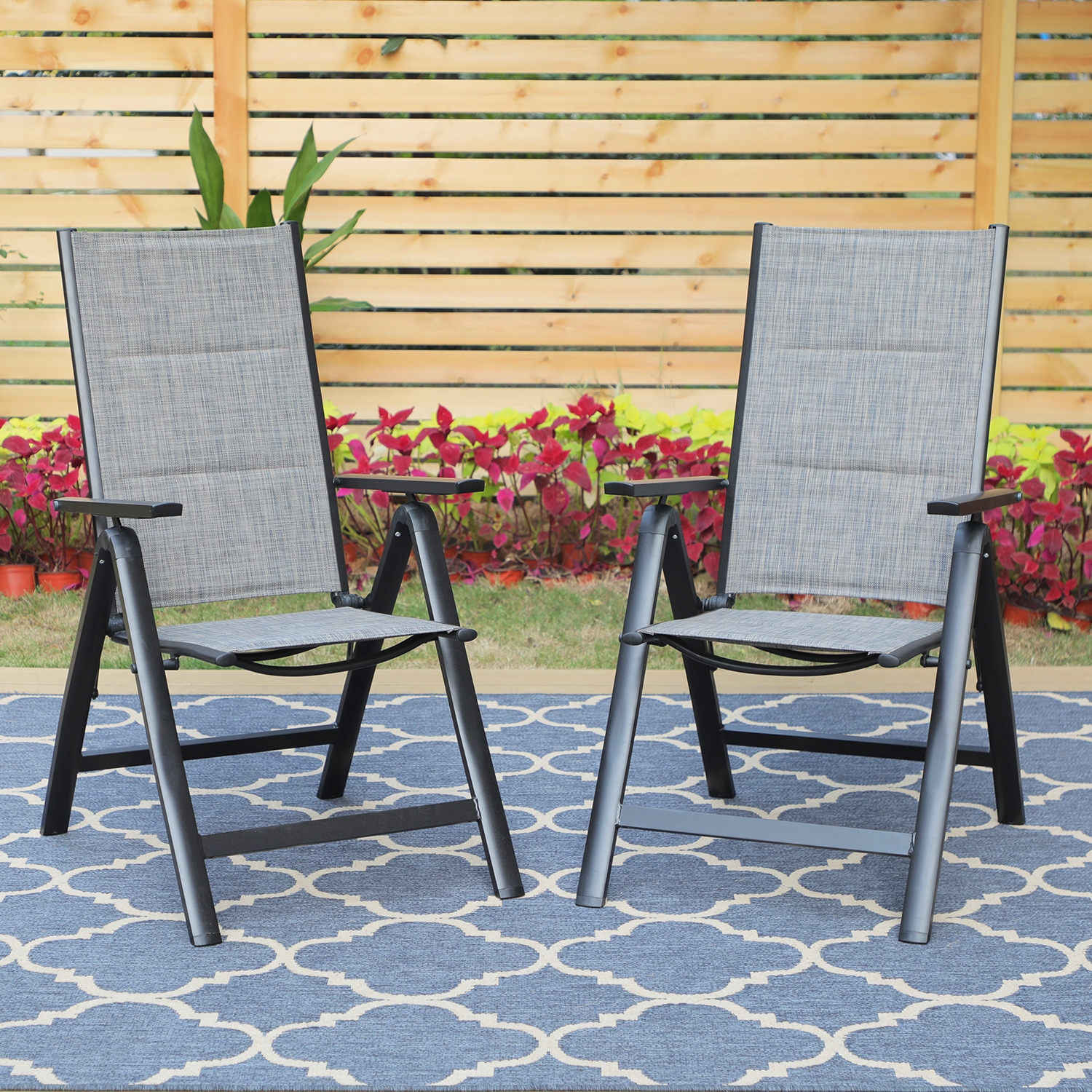 Two outdoor patio chairs with gray fabric seats and backs framed by black metal, set on a blue patterned outdoor rug, with a wooden fence and plants in the background.