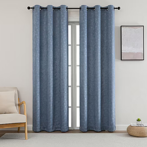 A pair of blue curtains hang on a rod in front of a window, accompanied by a beige cushioned chair, a small framed abstract artwork, and a round woven rug with a small plant on it.