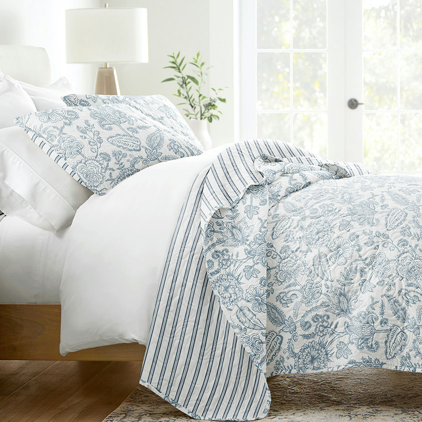 Bedding set with blue floral and striped patterns including a comforter, pillows, and shams in a bright room with a window.