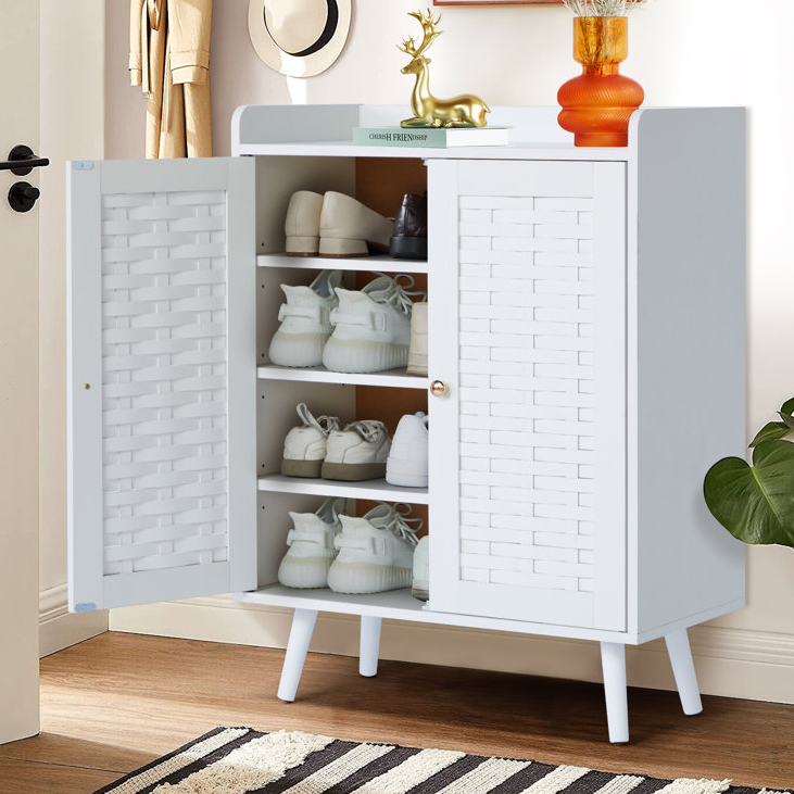 A white shoe storage cabinet with two slatted doors, four interior shelves, and raised legs, holding various shoes. An orange vase and decorative item are on top.