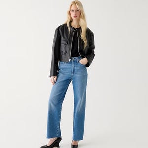 A woman is wearing high-rise slim wide-leg jeans in a classic blue denim wash with a visible button fly and traditional five-pocket styling. She is also wearing a black leather jacket and black heels.