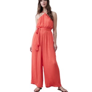 A woman is wearing a sleeveless, halter-neck, coral-colored jumpsuit with a pleated design and a waist tie. She pairs it with sandals and a metallic bangle.