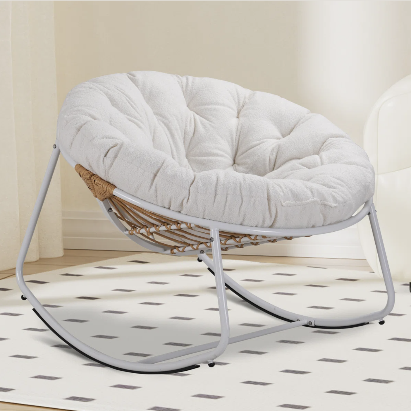 A round, cushioned rocking chair with a cozy white fabric and a metal frame, featuring a wicker design on the sides, positioned on a patterned rug indoors.