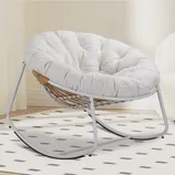 A round, cushioned rocking chair with a cozy white fabric and a metal frame, featuring a wicker design on the sides, positioned on a patterned rug indoors.