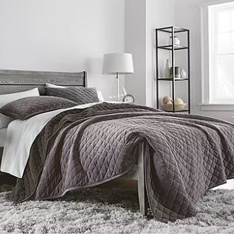 A gray upholstered bed with a quilted comforter set beside a metal frame shelf with decorative items.