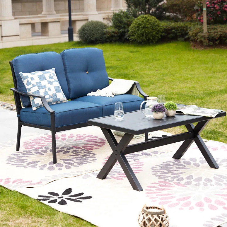 A 2-person patio set featuring a dark metal loveseat with blue cushions and armrests, accompanied by a matching rectangular coffee table. It rests on a decorative outdoor rug with a floral pattern, and includes accent items like glassware and small decorative plants.