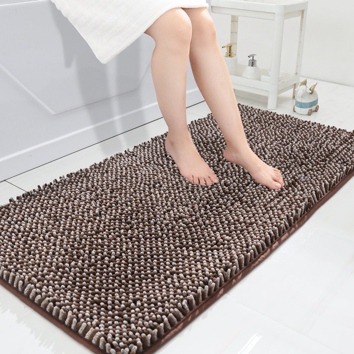 A person with bare legs sits on a bath mat with a shaggy, textured surface, placed on a white tile floor next to a bathtub and a small table holding toiletries.
