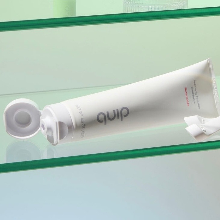 A tube of Quip Watermelon Anticavity Toothpaste with a white cap and a minimalist design on a glass shelf.
