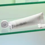 A tube of Quip Watermelon Anticavity Toothpaste with a white cap and a minimalist design on a glass shelf.