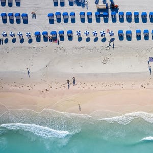 Aerial view of a beach with rows of blue beach loungers and matching umbrellas, and gentle waves reaching the shore.