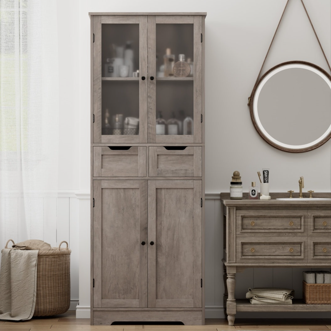 Rustic bathroom setup with a wooden linen cabinet featuring glass doors and drawers, alongside a matching vanity with a round mirror and various toiletries. A wicker basket with towels is placed nearby, on a wooden floor.