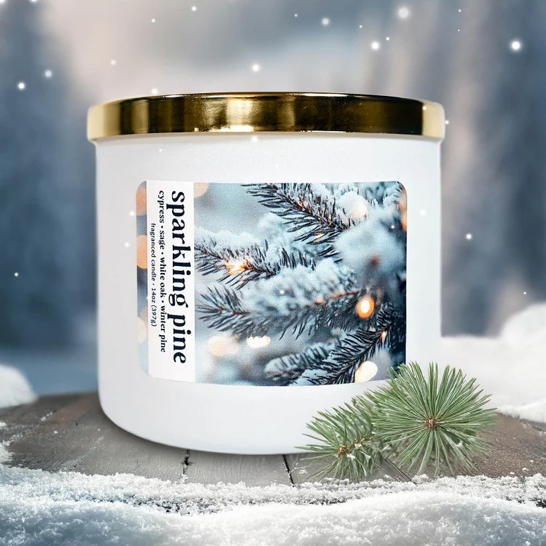 A white candle with a gold lid labeled \“sparkling pine\“ features a winter scene with snow-covered pine branches. Scents include cypress sage, white oak, and blue spruce.
