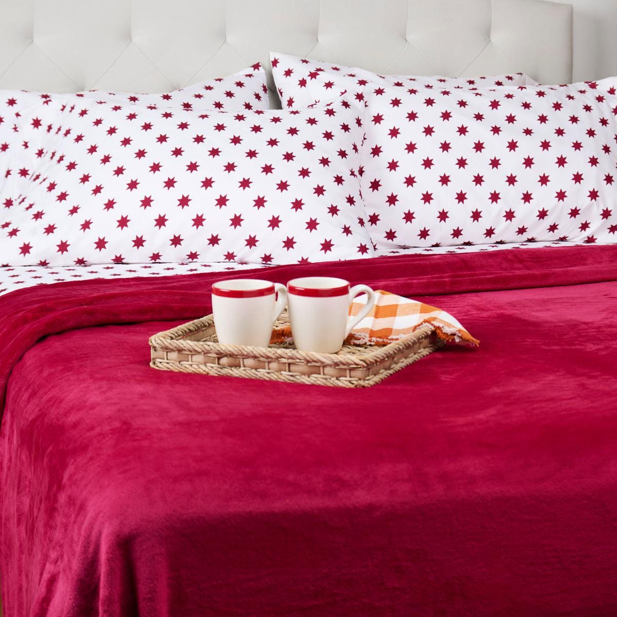 Red star-patterned bed linens with a matching blanket, accompanied by two white mugs with red rims on a woven tray, and an orange checkered napkin.