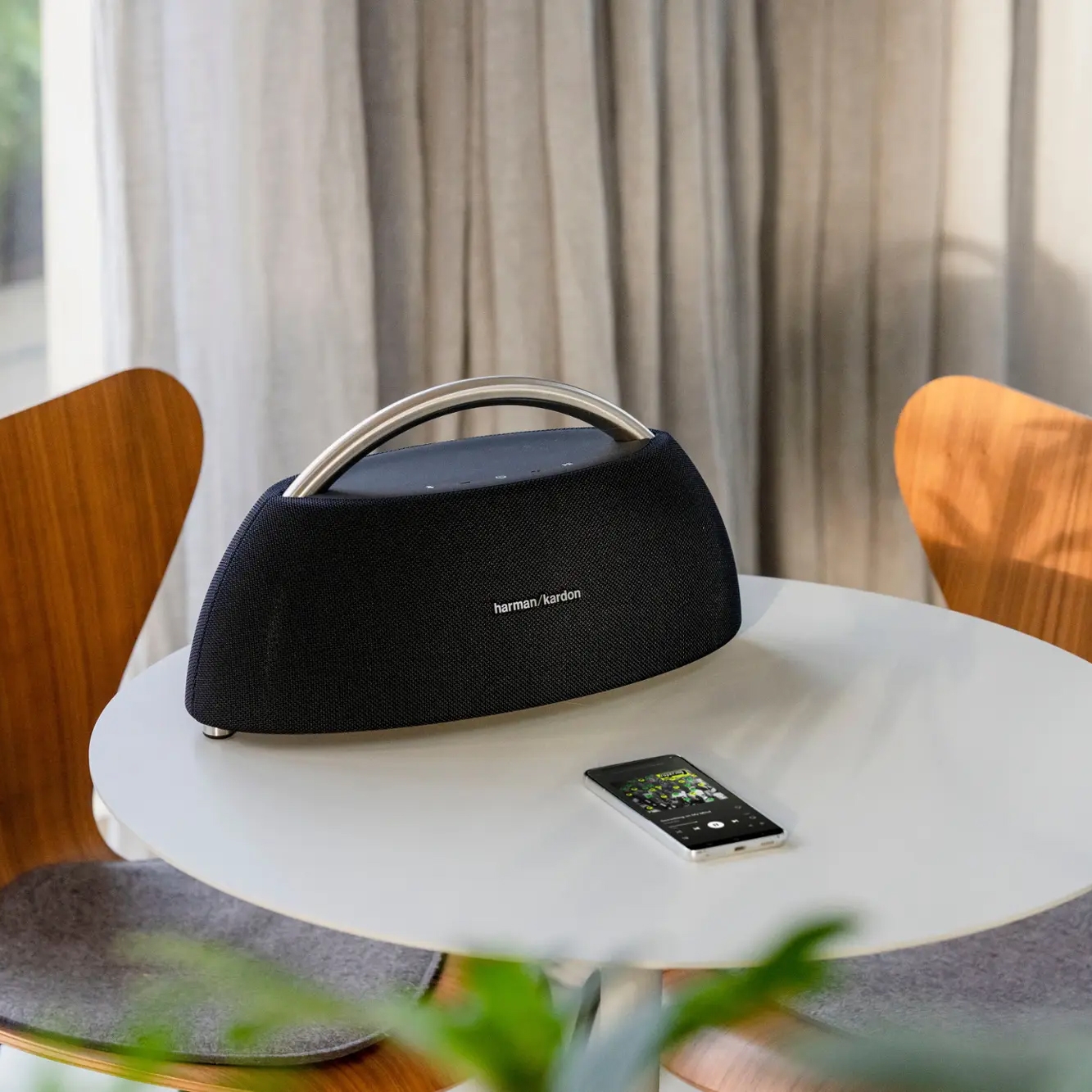 A Harman Kardon speaker with a metallic handle is placed on a round table next to a smartphone, surrounded by modern wooden chairs and curtains.