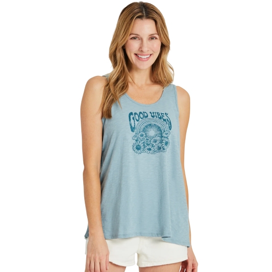 A woman is wearing a light blue sleeveless top with a \“Good Vibes\“ graphic and white shorts. She has long, wavy hair and is smiling.