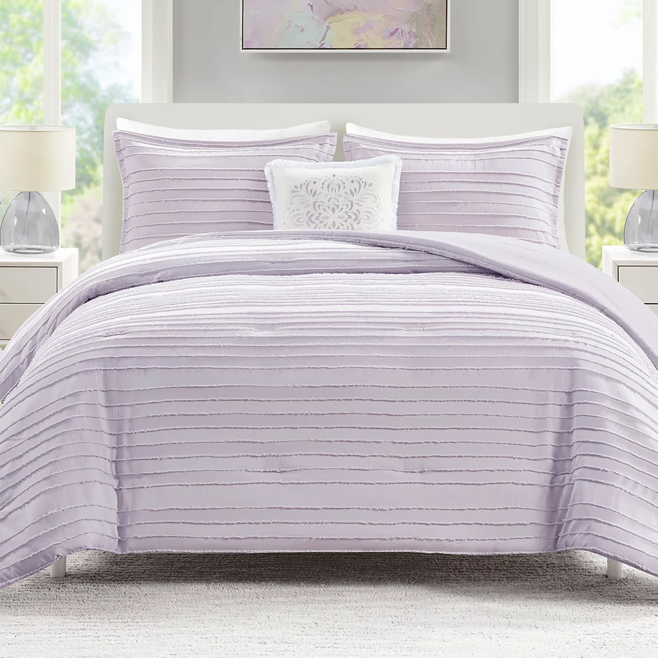 The JLA Home Ottie Comforter Set features a lavender comforter with horizontal textured lines, accompanied by matching pillow shams and a decorative white accent pillow with a floral design.