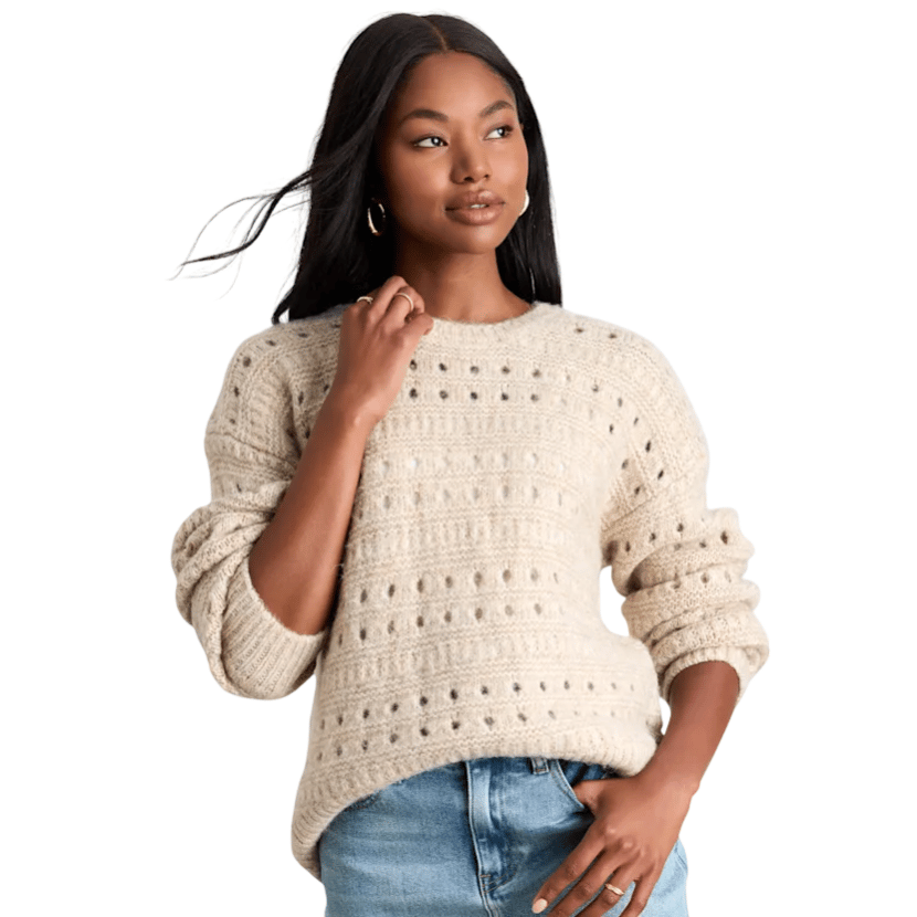 A woman is wearing a textured, beige knit sweater with a perforated design and light blue jeans.