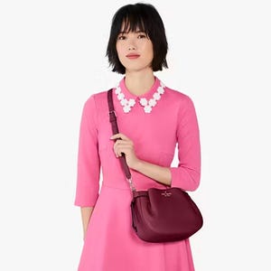 A woman is wearing a pink dress with a white floral necklace and carrying a burgundy Kate Spade Dumpling Crossbody bag with an adjustable shoulder strap.