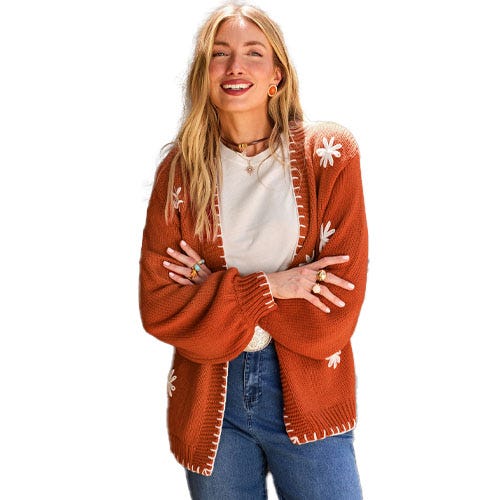 A woman is wearing a rust-colored cardigan with white floral embroidery, a cream shirt, and blue jeans.