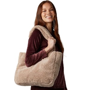 A person is carrying a large tan, fluffy tote bag over their shoulder while wearing a long-sleeved, dark red outfit.
