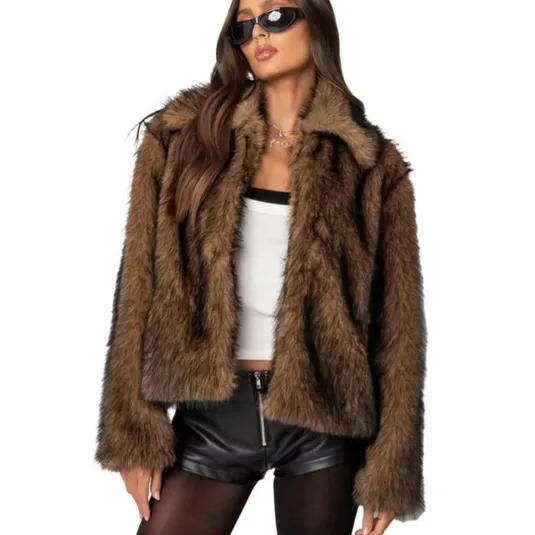 A woman in a brown faux fur jacket, white cropped top, black leather shorts with a front zip, black tights, sunglasses, and a necklace.