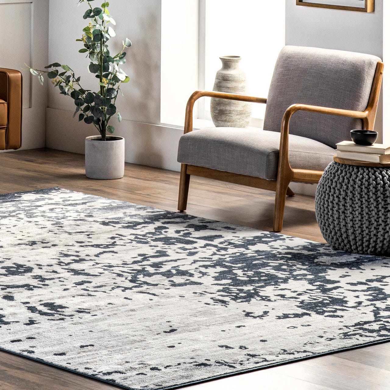 A modern living room features a gray upholstered chair with wooden arms, a patterned area rug, a gray knit pouf, a potted plant, and a ceramic vase on a wooden floor.