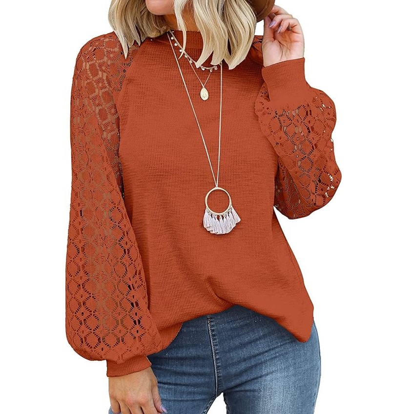 A woman is wearing a rust-colored blouse with lace sleeves and a layered necklace with a round pendant and a fringe detail.