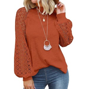 A woman is wearing a rust-colored blouse with lace sleeves and a layered necklace with a round pendant and a fringe detail.
