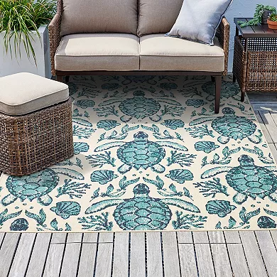 An outdoor area rug with a sea turtle design is placed on a wooden deck, accompanied by a beige cushioned sofa and a rattan side table.