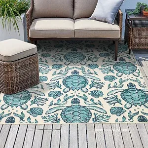 An outdoor area rug with a sea turtle design is placed on a wooden deck, accompanied by a beige cushioned sofa and a rattan side table.