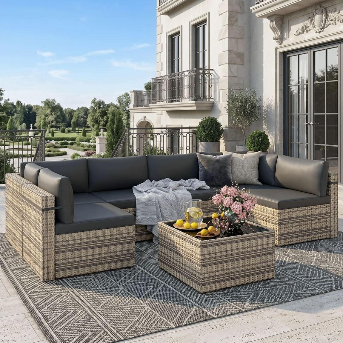 Outdoor patio set with gray cushions on a woven rattan sectional sofa and matching coffee table, featuring a tray with lemonade and pink flowers. The setting includes a decorative gray geometric-patterned rug on a spacious terrace.