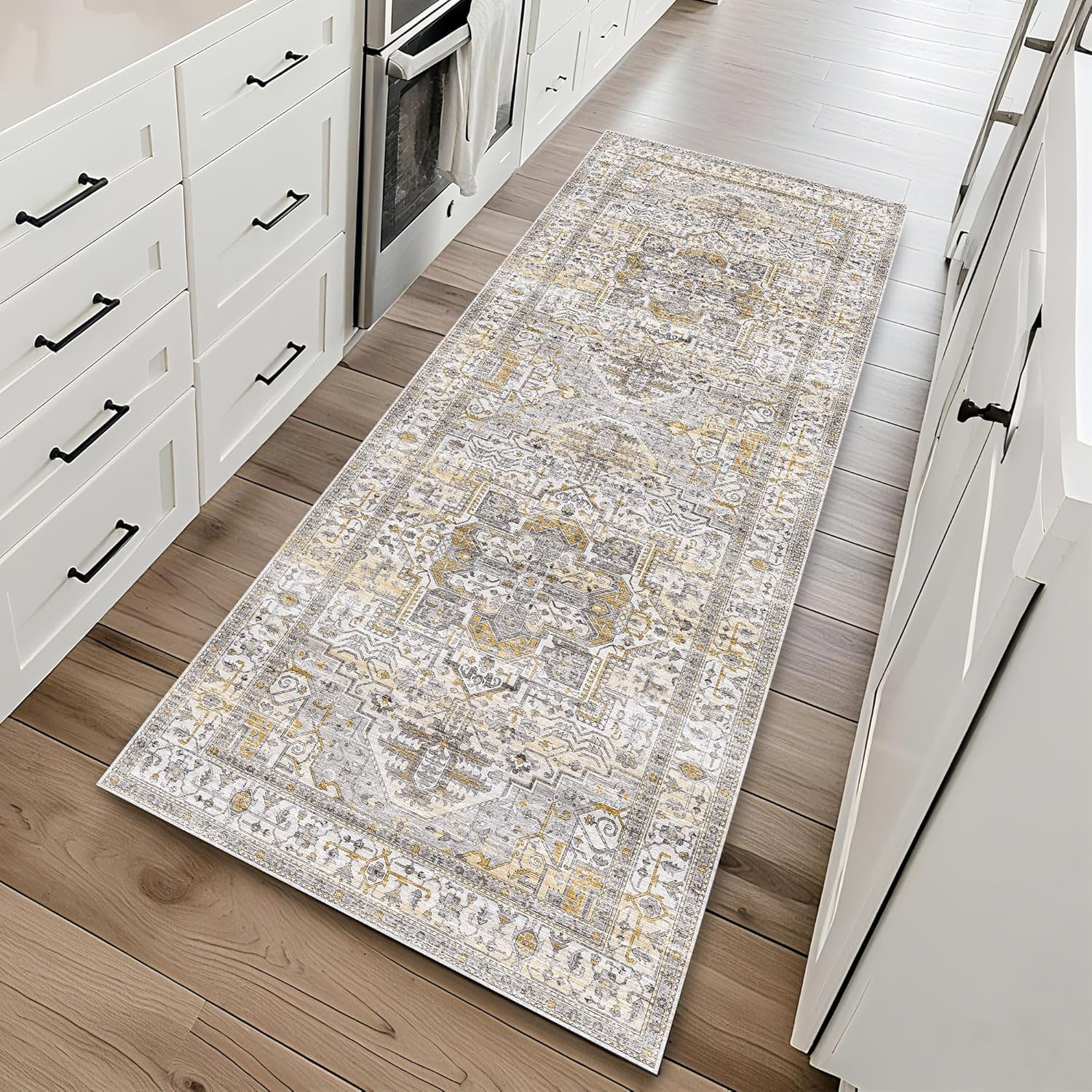 A patterned runner rug with a vintage design featuring grey, gold, and beige tones placed on a light wood floor in a kitchen setting with white cabinetry.
