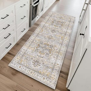 A patterned runner rug with a vintage design featuring grey, gold, and beige tones placed on a light wood floor in a kitchen setting with white cabinetry.