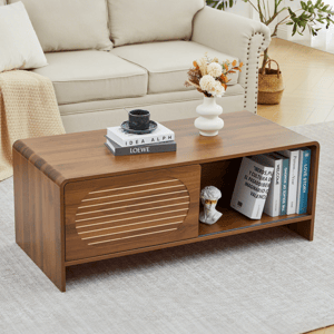 A wooden coffee table with a textured sliding door, displaying books, a vase with flowers, and a cup. It is placed in front of a beige couch on a light rug, with a leafy plant in the background.