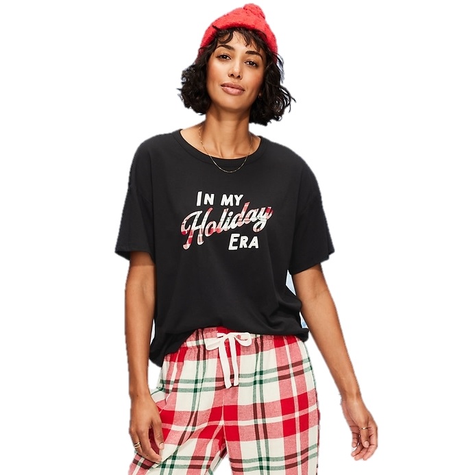 A woman wearing a black T-shirt with \“In my Holiday Era\“ text, plaid pajama pants in red, white, and green, and a red beanie.