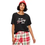 A woman wearing a black T-shirt with \“In my Holiday Era\“ text, plaid pajama pants in red, white, and green, and a red beanie.