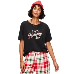 A woman wearing a black T-shirt with \“In my Holiday Era\“ text, plaid pajama pants in red, white, and green, and a red beanie.