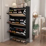 A black shoe rack with multiple tilt-out compartments displays various shoes. It's placed in a well-lit entryway, accompanied by decorative items like a lamp and plant.