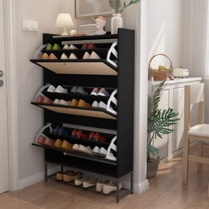A black shoe rack with multiple tilt-out compartments displays various shoes. It's placed in a well-lit entryway, accompanied by decorative items like a lamp and plant.