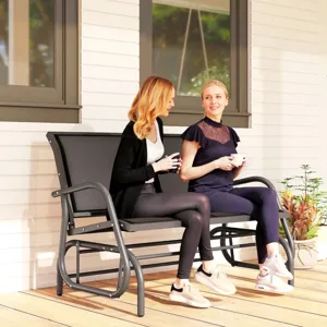 Two women sit on a metal outdoor glider bench, each holding a cup, on a wooden porch with a white wall background. A small potted plant sits beside them.
