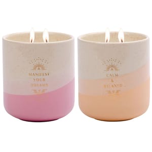 Two ceramic jar candles with double wicks, one pink with \“Manifest Your Dreams,\“ the other peach with \“Calm & Relaxed\“ in gold lettering, featuring a smooth two-tone design.