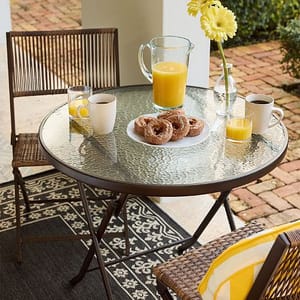 Round glass-top patio table with two wicker chairs, featuring mugs, a glass of orange juice, a pitcher with juice, a plate of donuts, and a vase with yellow flowers on a patterned outdoor rug.
